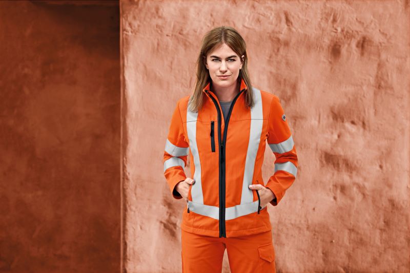 Une femme porte des vêtements de signalisation orange et a les mains dans les poches.